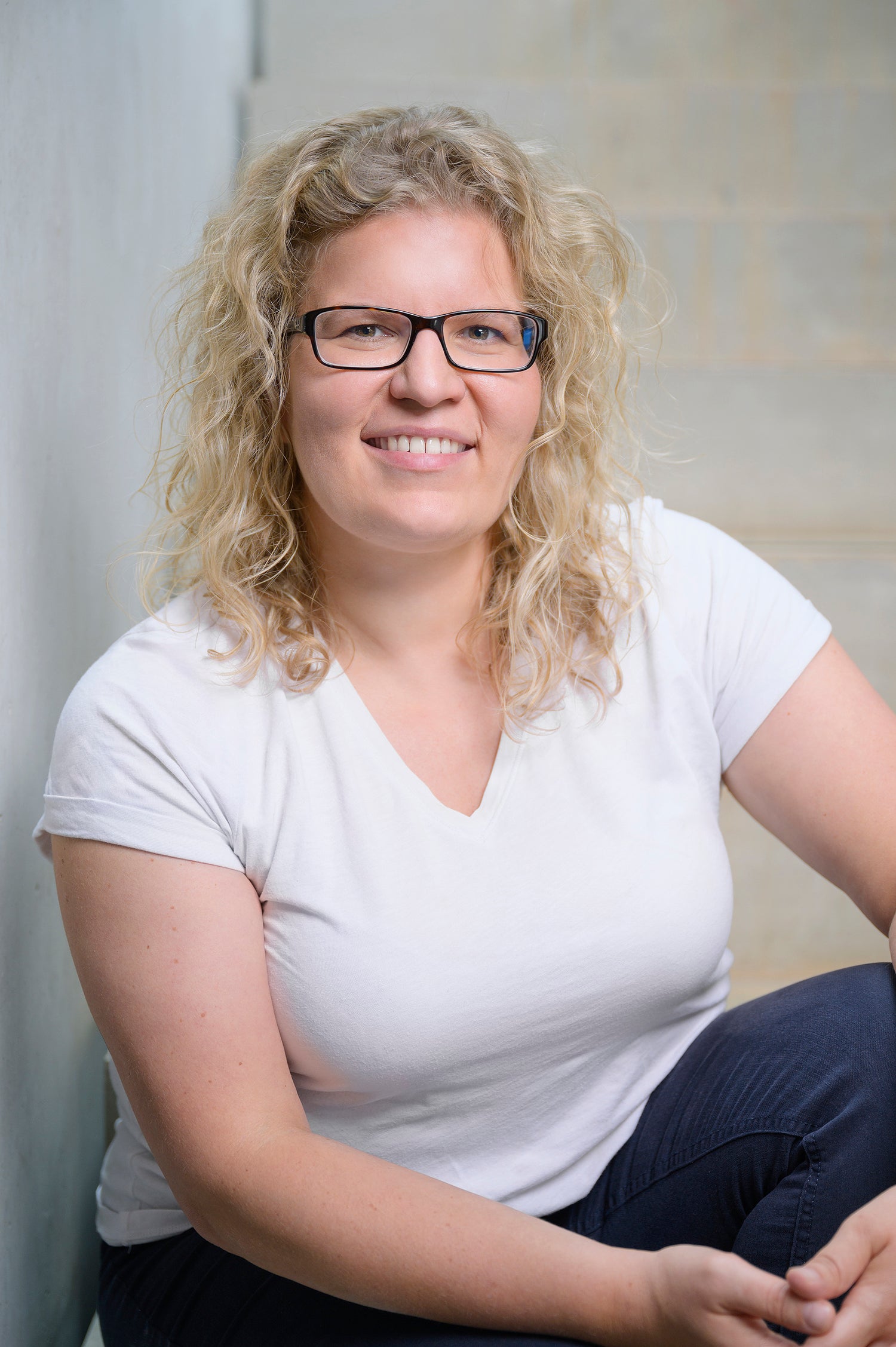 Porträt einer blonden, lockigen Frau mit schwarzer Brille und weißem Oberteil.